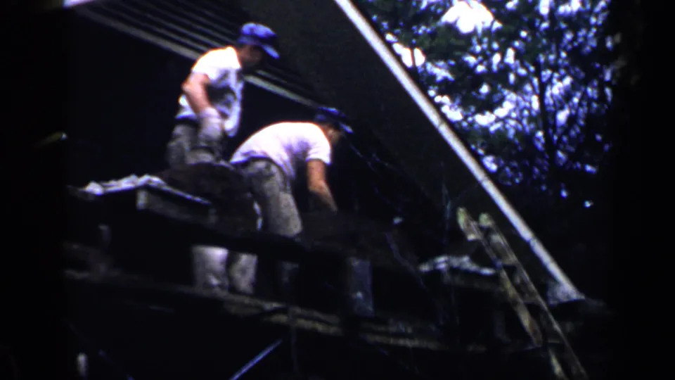 Two adult men in caps continue roof work in the 1957 Birmingham 8mm footage, seen from below with trees behind the structure.