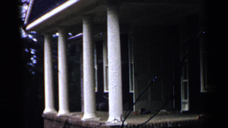 The final frame holds on the Birmingham house porch, with white columns, dark entryway, brick base, and foreground branches.