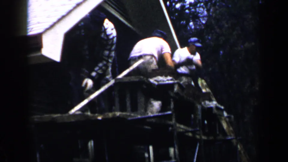 1957 home-movie frame of workers on an elevated Birmingham scaffold, with brickwork, caps, and trees visible.