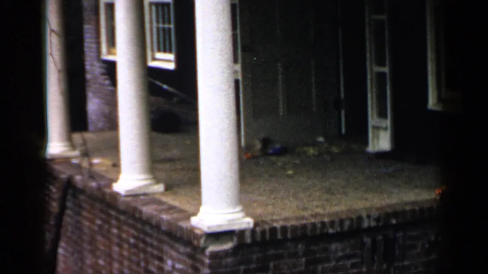 1957 Birmingham home-movie frame showing a brick house porch with white columns, dark doorway, windows, and scattered objects near the entrance.