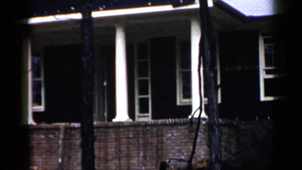 1957 Birmingham home-movie frame with a wider porch view, white columns, brick railing, dark windows, and tree trunks crossing the foreground.