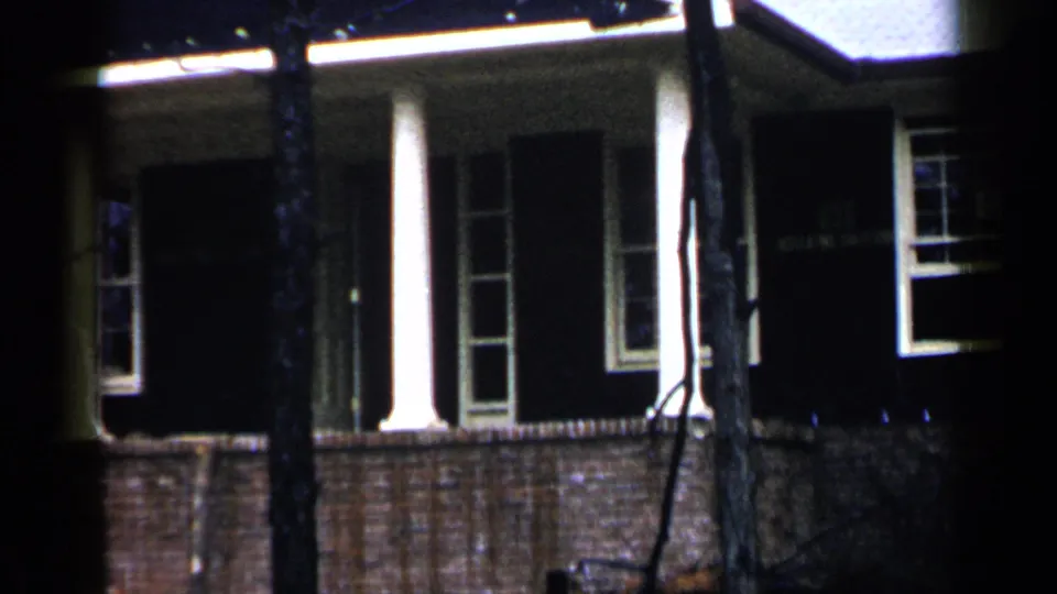 1957 Birmingham home-movie frame showing a brick residential exterior at night, partly obscured by tree trunks.