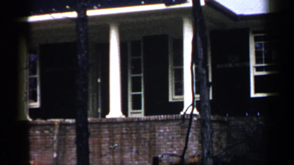 1957 Birmingham frame of a dark brick house exterior with white columns and windows seen through foreground trees.