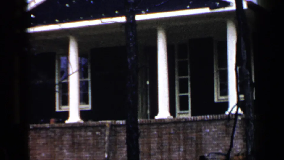 Final 1957 Birmingham frame showing the brick residential facade, columns, shutters, and foreground tree trunks.