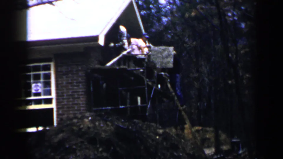 Distant workers continue exterior construction or repair beside a brick Birmingham house in 1957 8mm footage.