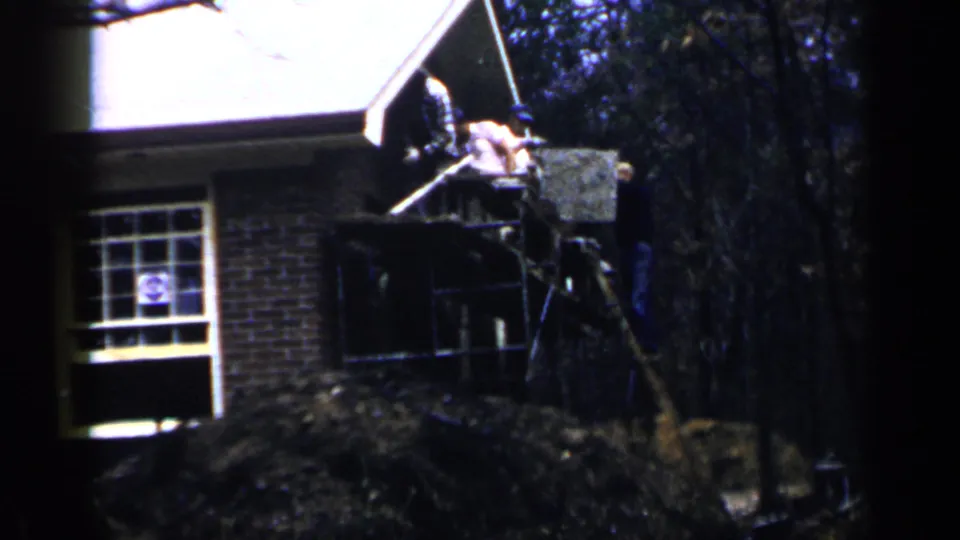 1957 Birmingham home-movie frame showing men on scaffolding beside a brick house during porch construction.