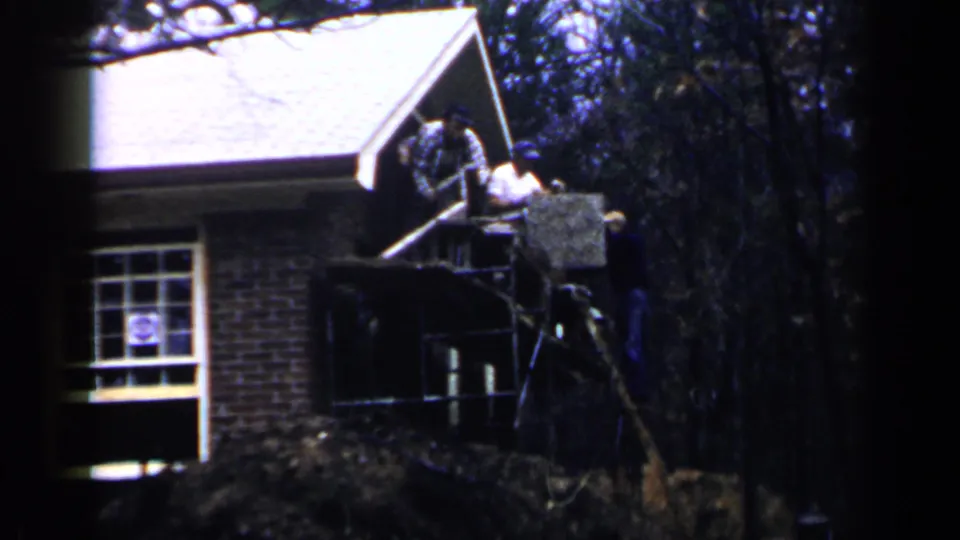 1957 Birmingham home-movie frame with workers on porch scaffolding beside a brick house and wooded background.