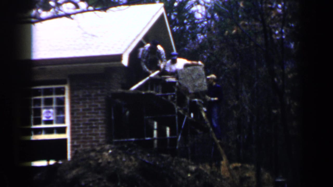 Workers on scaffolding beside a brick house with trees in the background.