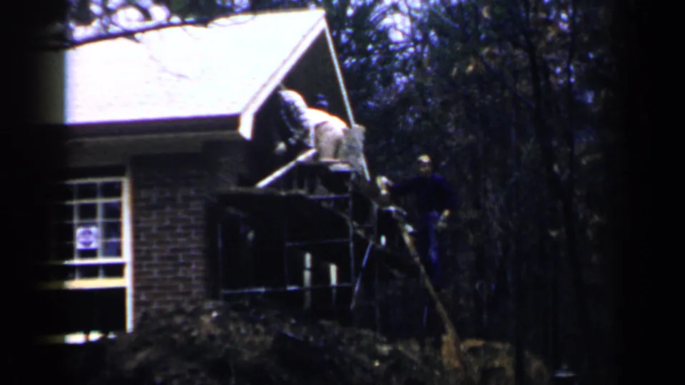 Workers appear on scaffolding at a Birmingham brick residence, with one figure near a large block or panel by the roofline.