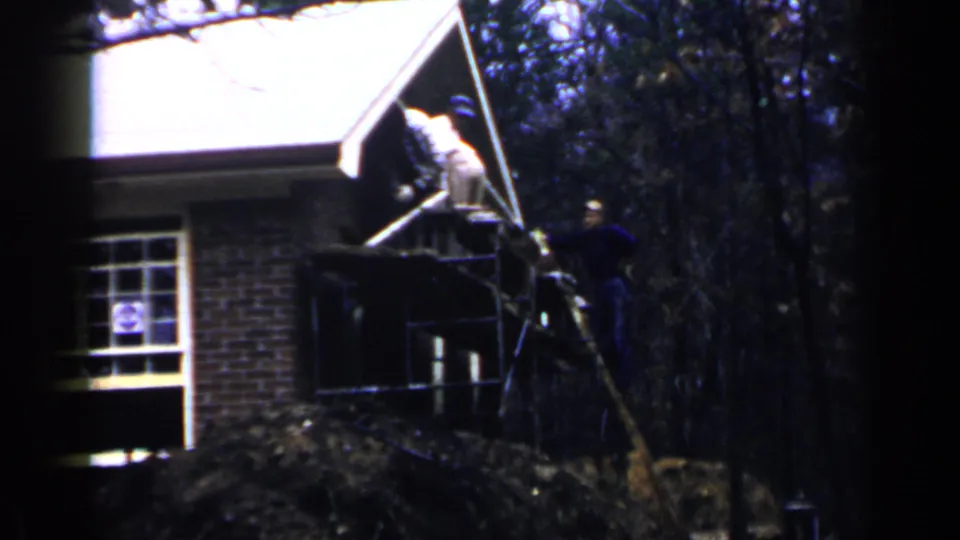 1957 Birmingham footage frame with workers on and around scaffolding beside a wooded residential brick house.