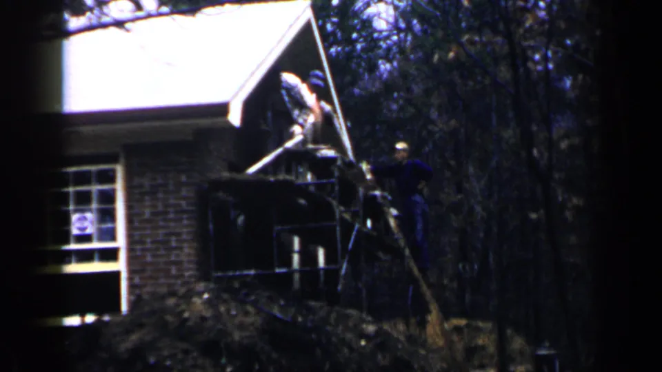 1957 Birmingham home-movie frame showing men on a scaffold at a brick house porch, with a window, roof gable, and wooded background.