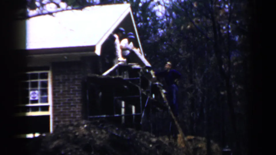 1957 Birmingham frame showing workers clustered at a porch opening on a brick house, with a scaffold and wooded yard visible.