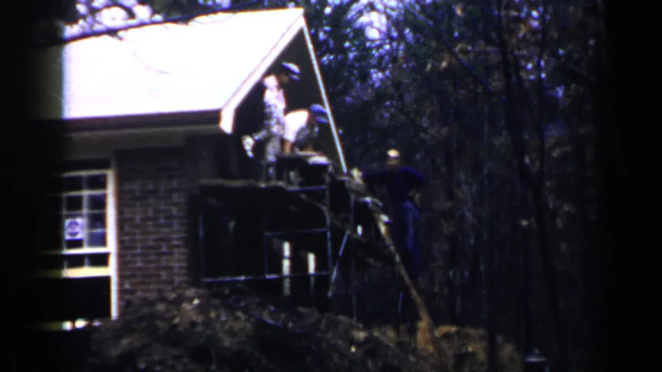 1957 Birmingham home-movie frame with men positioned on a scaffold and near a brick house porch during exterior construction or repair.