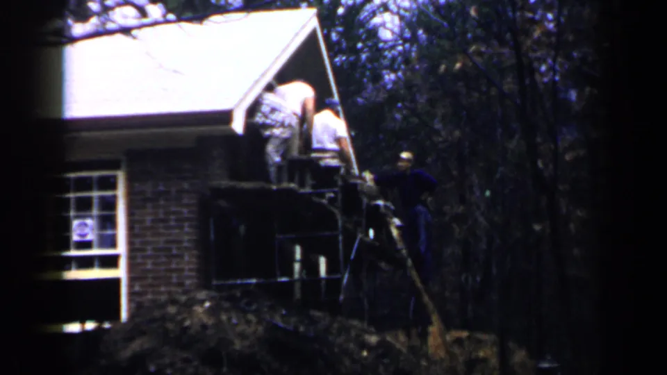 1957 Birmingham frame showing men bent toward a porch opening from a scaffold, with brickwork, window, roofline, and trees visible.