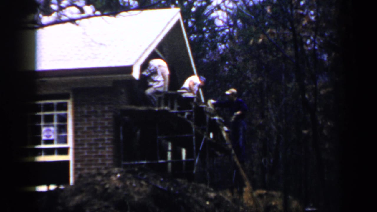 Two men work on scaffolding beside a brick house under trees.