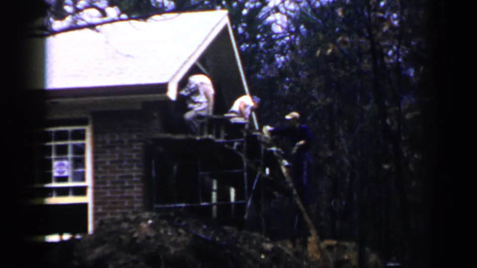 1957 Birmingham home-movie frame showing two men on scaffolding near the roof edge of a brick house surrounded by trees.
