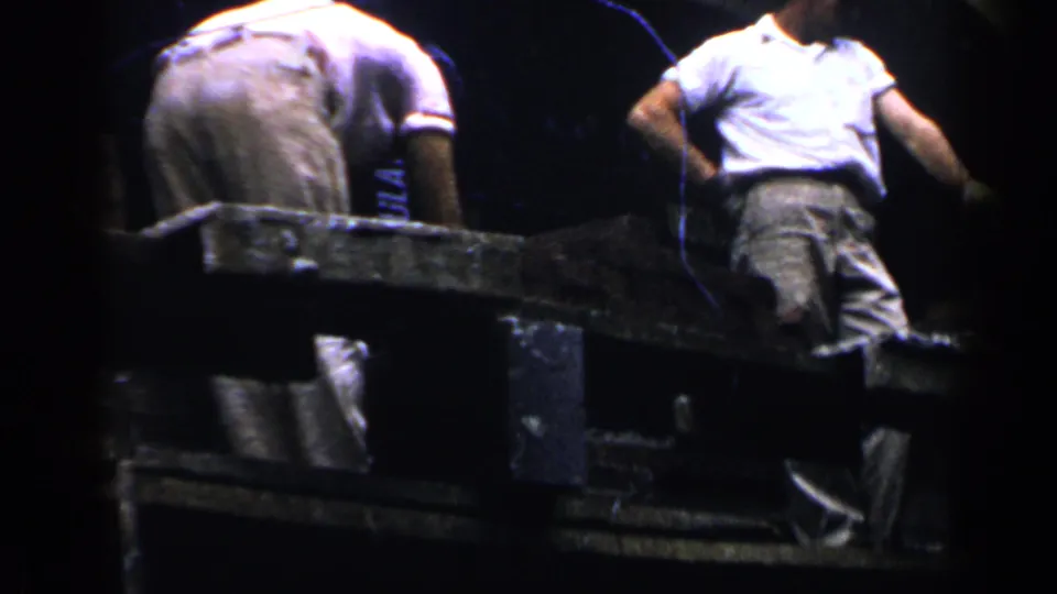 Two adult men work from a scaffold in a dark, close 8mm frame, one bending forward while another stands in a white shirt and cap.