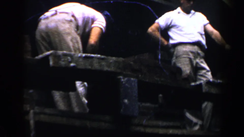 Close home-movie view of two men on scaffolding during roof or exterior house work, with heavy shadow and soft 8mm color.