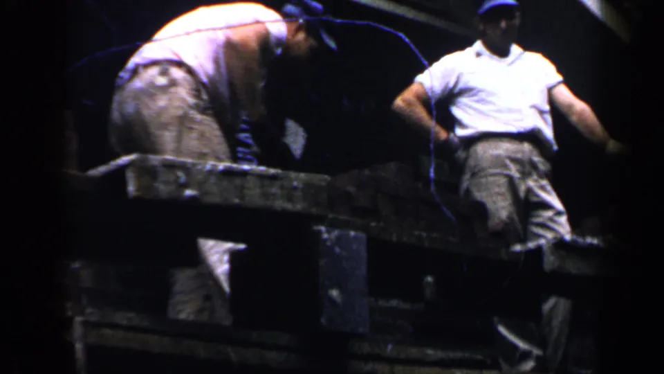 Late frame from the 1957 Birmingham clip showing two men on scaffold planks, one bent over and one standing in a white shirt near the house roof.