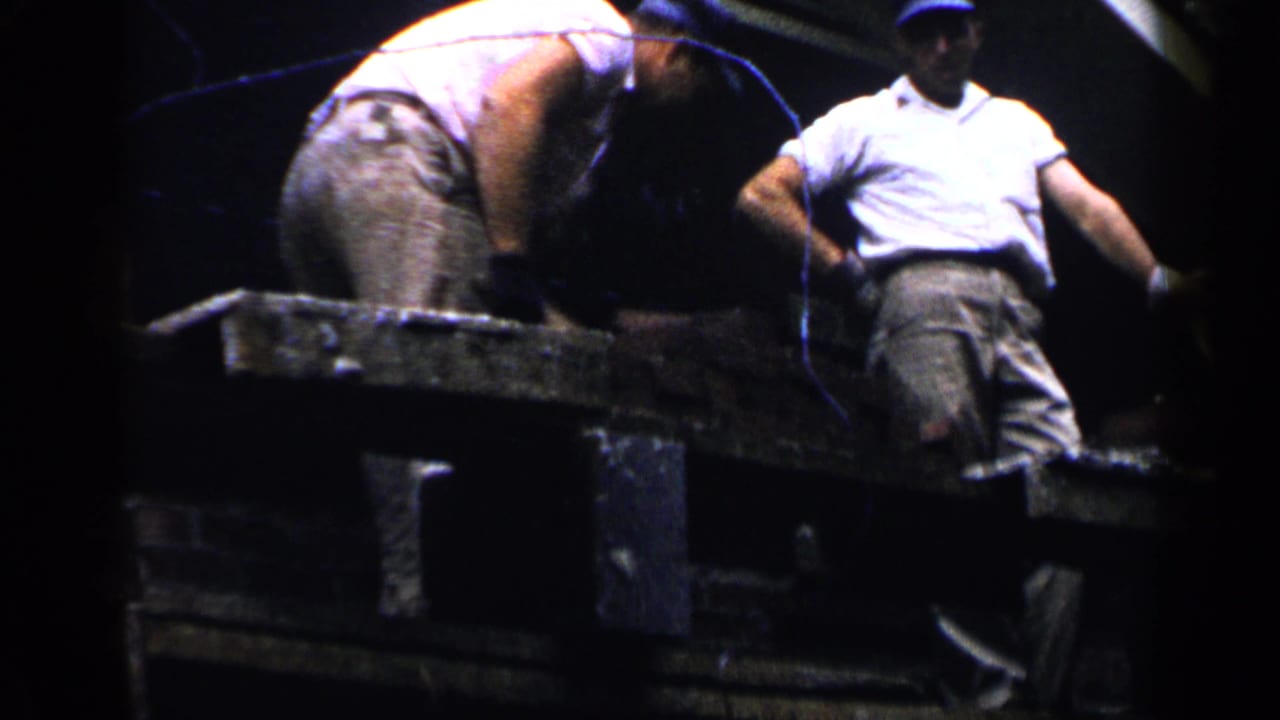 Two construction workers stand near a brick wall and work platform in strong sunlight.