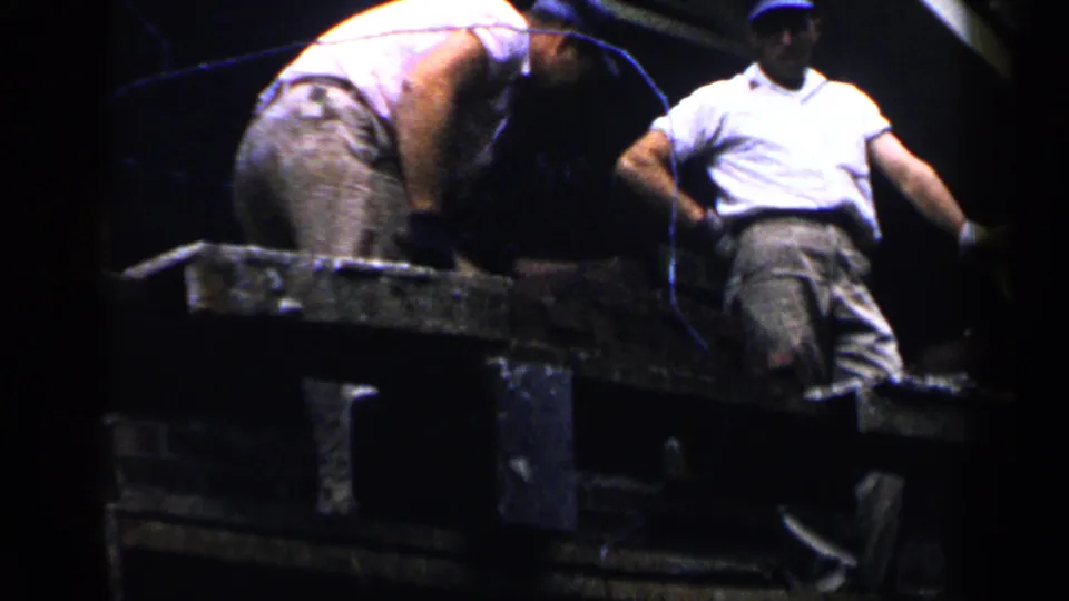 1957 Birmingham 8mm frame showing two construction workers near a brick wall, with one bent over a platform and another standing nearby.