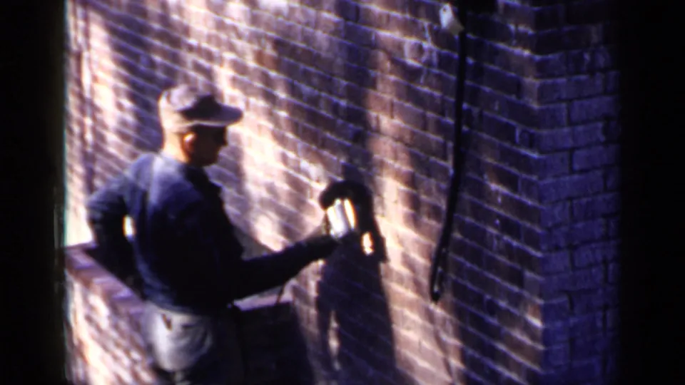 1957 Birmingham frame of a construction worker using a handheld power tool on a brick wall beside a shadowed corner.