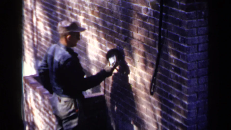 1957 Birmingham 8mm frame with a worker in dark clothing holding a tool against a brick wall in bright sunlight and heavy shadow.