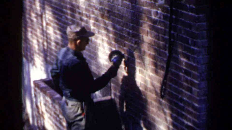 1957 Birmingham frame showing a capped construction worker using a tool on a brick wall, with strong light and shadow across the masonry.