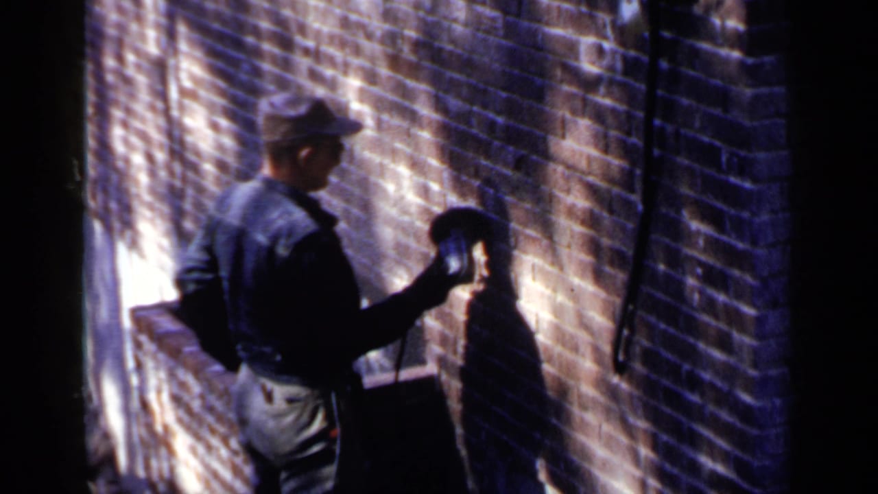 Man in a cap holds a power tool against a sunlit brick wall.
