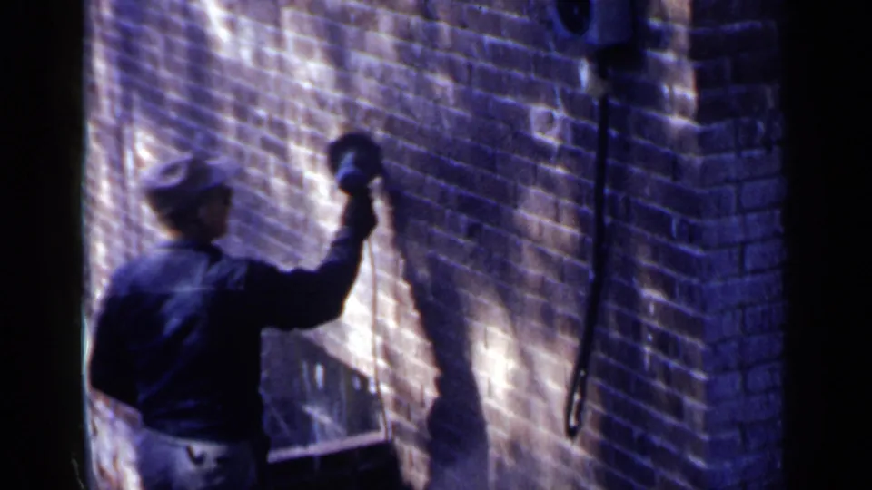 The worker stands close to the Birmingham brick wall, tool raised, with strong sunlight and shadow defining the work area.