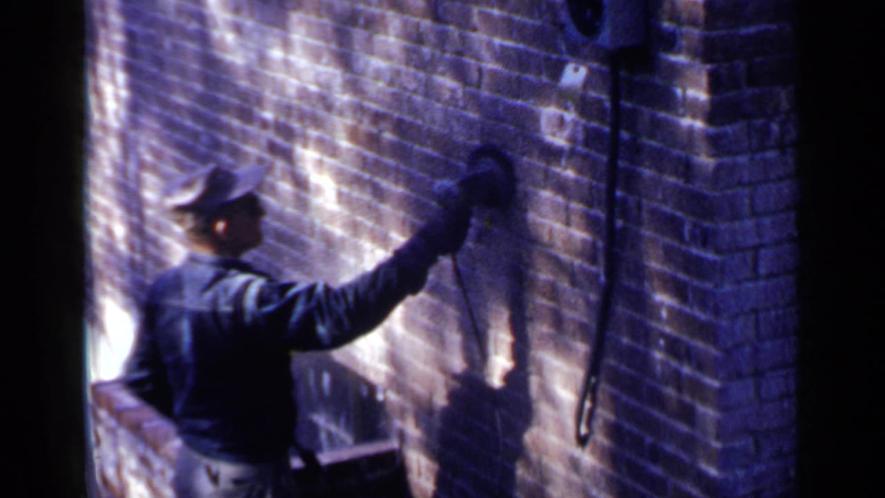 Man in cap works a handheld tool against a sunlit brick wall.