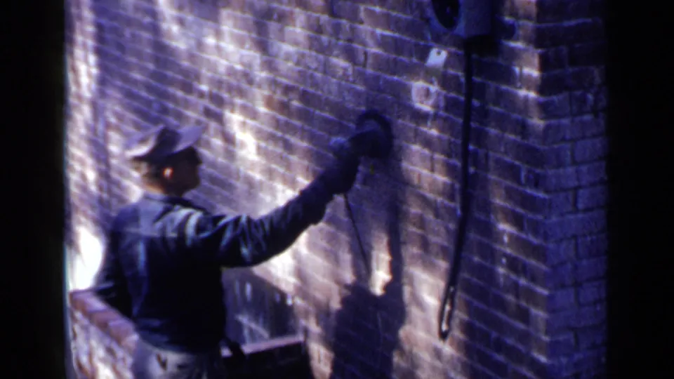 In 1957 Birmingham, Alabama, a man in a cap and dark jacket works a handheld tool on a brick exterior wall.