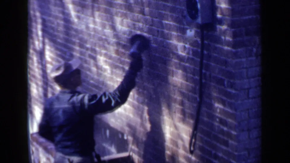 A 1957 Birmingham frame shows the man continuing work on the brick wall, filmed in soft 8mm color with deep shadow.