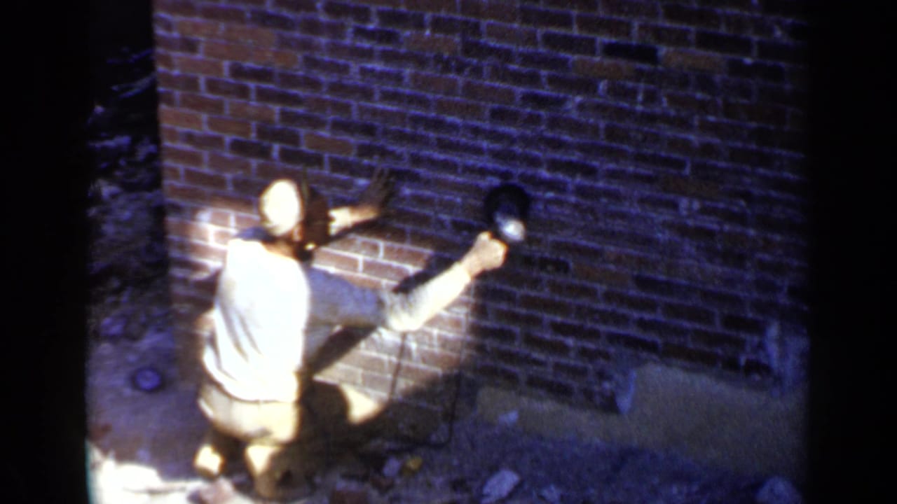 Man in a light shirt and cap sands a red brick wall with a power tool.