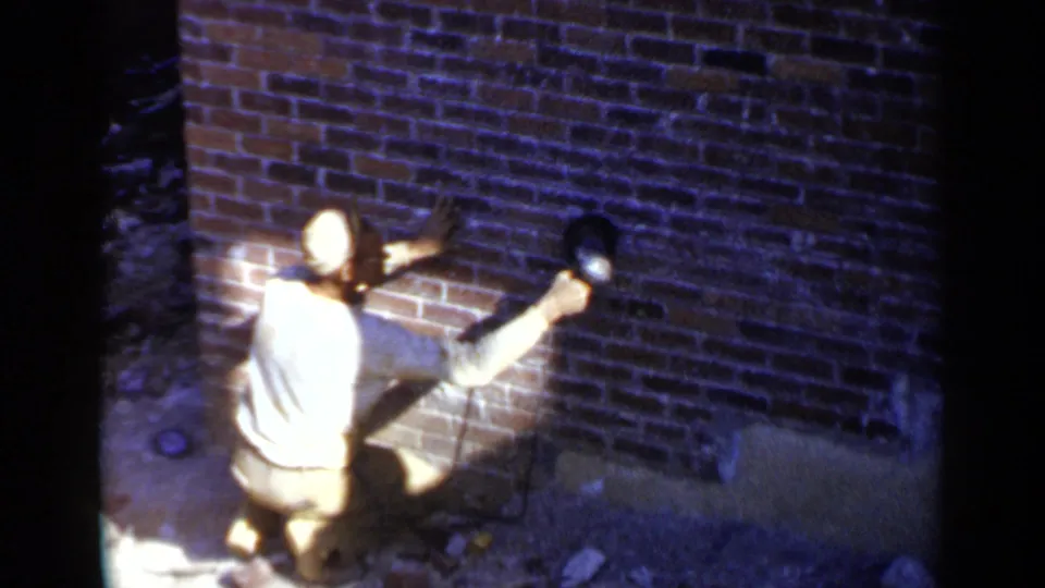 In 1957 Birmingham footage, a man kneels beside a red brick wall and presses a power sander against the masonry.