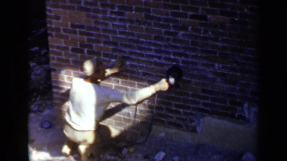 The worker extends one arm to hold a power sander against the red brick wall, with bright sunlight and dark shadow around the work area.