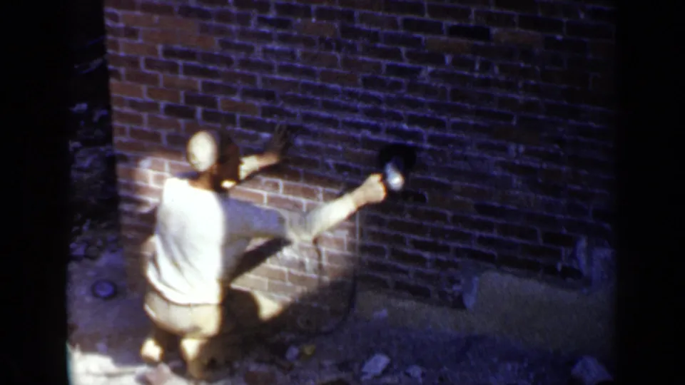 A man in a cap kneels close to the red brick wall, using a corded sander near the lower courses of masonry.