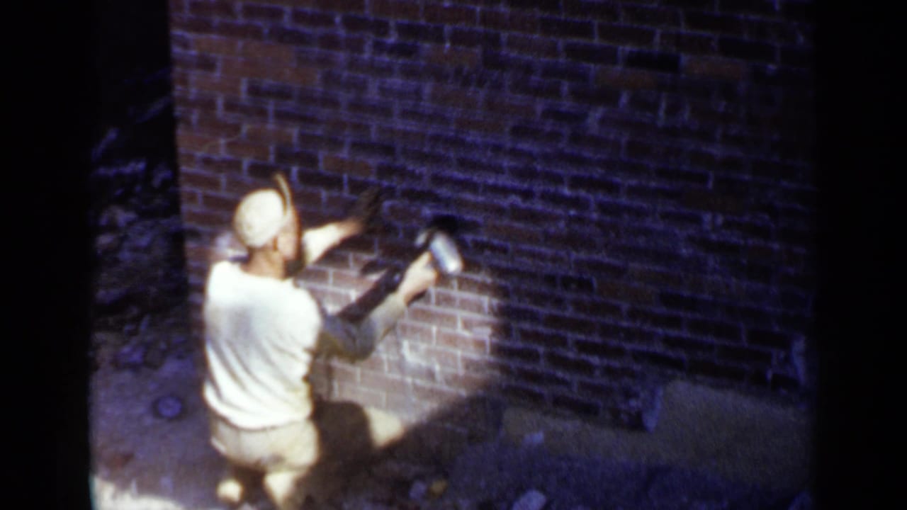 Worker in a cap kneels beside a brick wall using a handheld grinder.