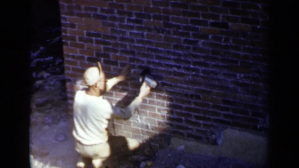 A worker in a light shirt and cap works close to a brick wall with a handheld grinder in the 1957 Birmingham footage.