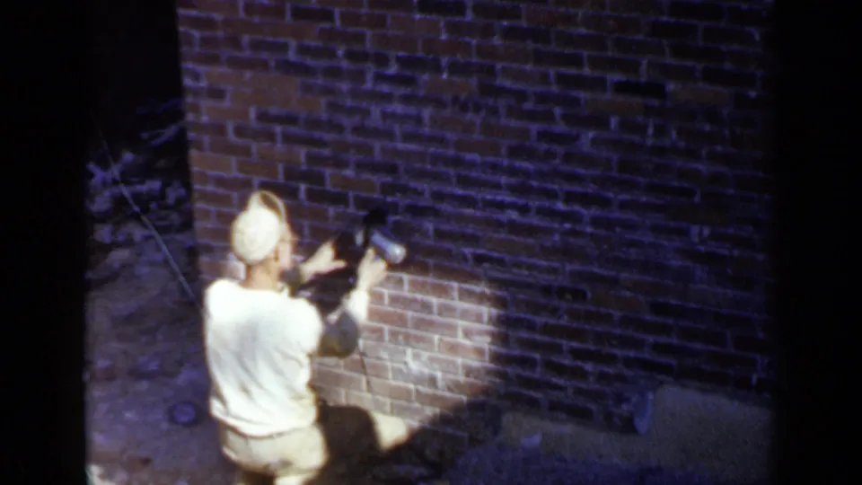 The 8mm frame shows a kneeling worker at the base of a brick wall, focused on lower brick cleaning or repair work.