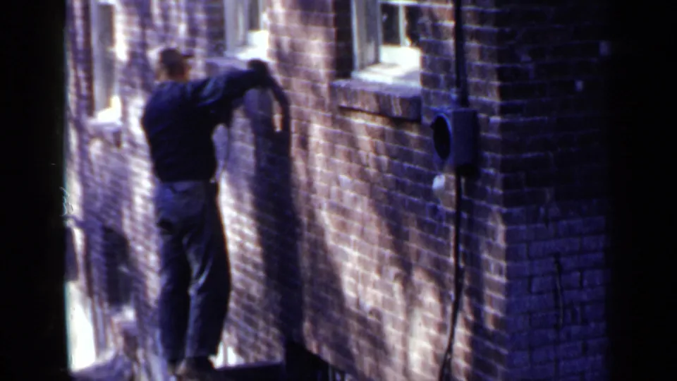 A second worker in a dark shirt appears to work on a higher brick-wall section near windows in the Birmingham 1957 footage.