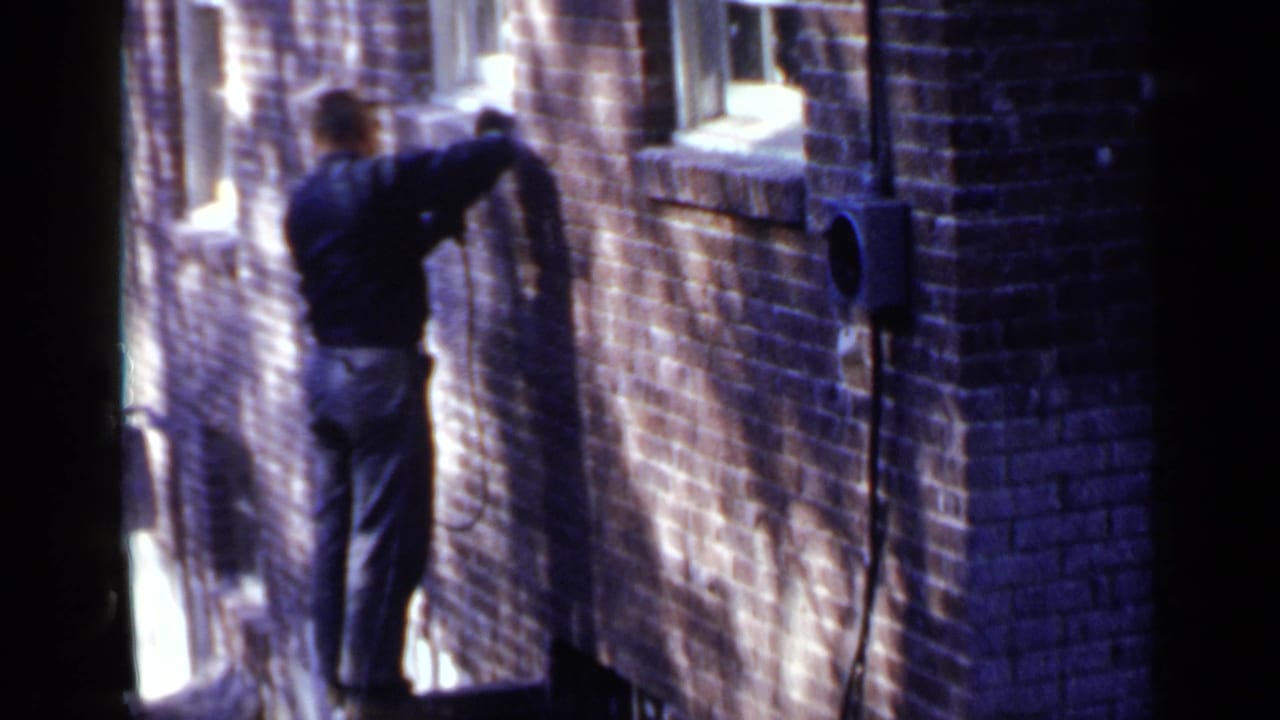 In 1957 Birmingham, a man in a hat stands near a brick building exterior while using a power tool on the wall.
