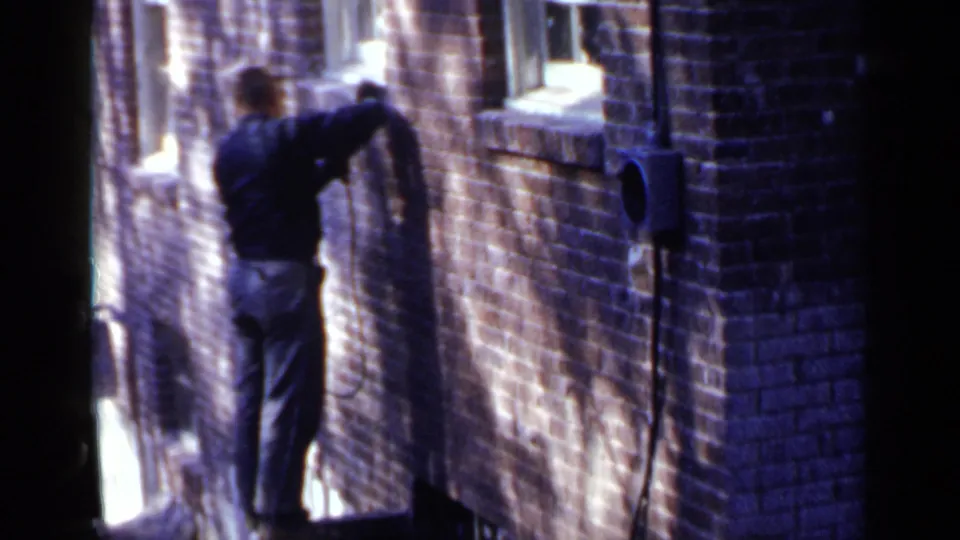 In 1957 Birmingham, a man in a hat stands near a brick building exterior while using a power tool on the wall.