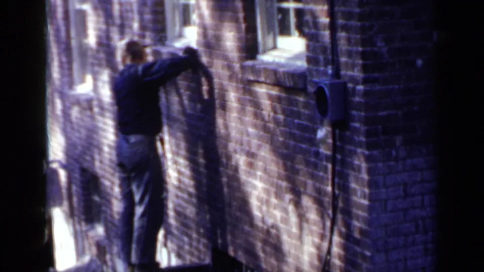 A man in work clothes faces the brick building wall, with the tool cord hanging nearby and bright sunlight falling across the surface.