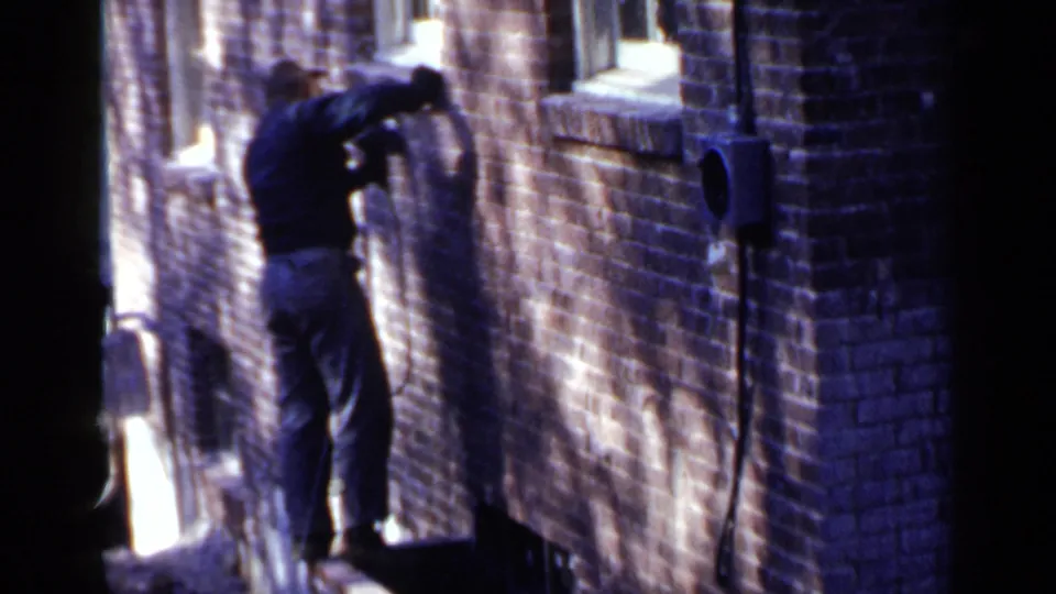 The Birmingham home-movie frame shows the man leaning into the brick exterior as he continues work with a corded tool near a window.