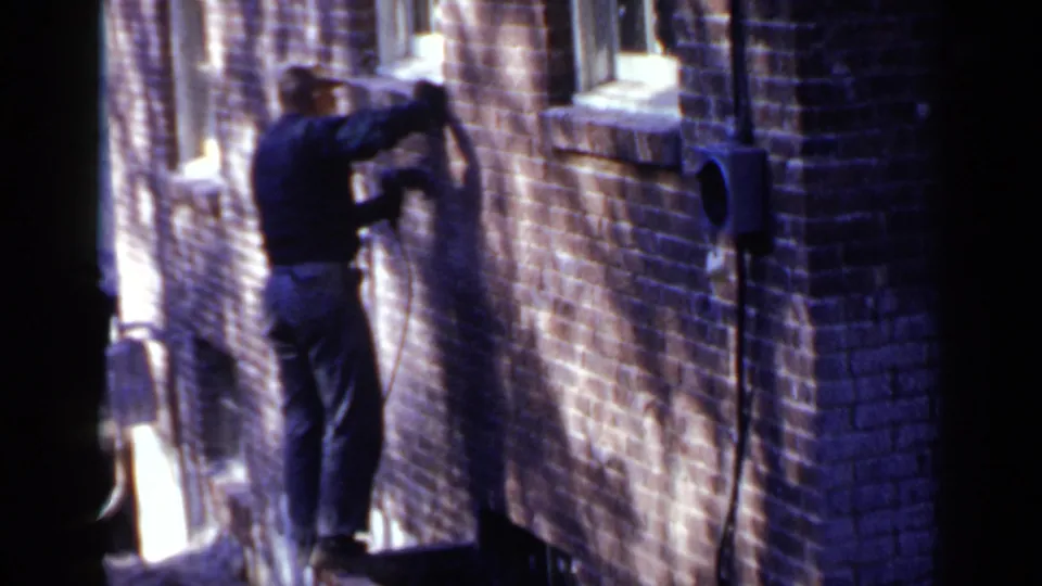 In 1957 Birmingham, a man in a dark jacket and cap appears to work against a sunlit brick wall in a narrow alley.