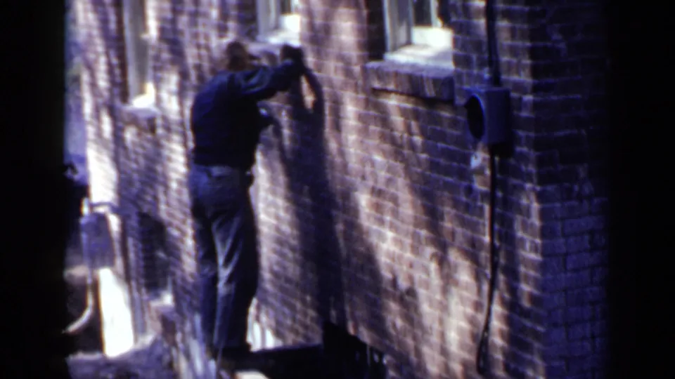 The man remains close to the Birmingham brick building, arm raised as if cleaning or inspecting the wall in the alley.
