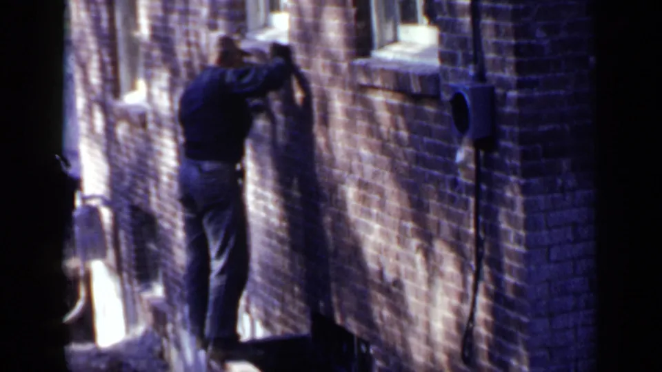 A candid 8mm frame shows the man leaning toward the brick building, with sharp sunlight and shadow crossing the alley wall.
