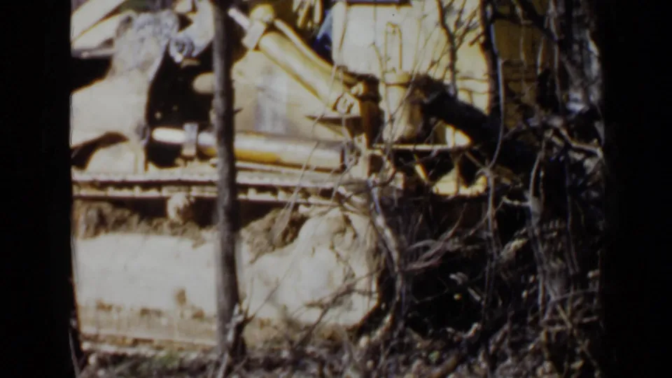 The frame shows a closer 1957 Birmingham view of bulldozer machinery obscured by trunks, brush, and uneven ground.
