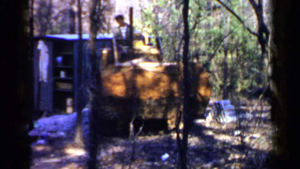 A man on a yellow bulldozer moves past a small blue shed in the wooded 1957 Birmingham footage.